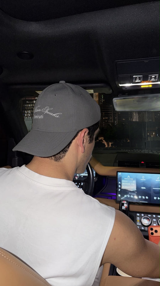 A man wearing a gray baseball cap with white script embroidery and a white sleeveless shirt, seen from behind inside a car cabin.